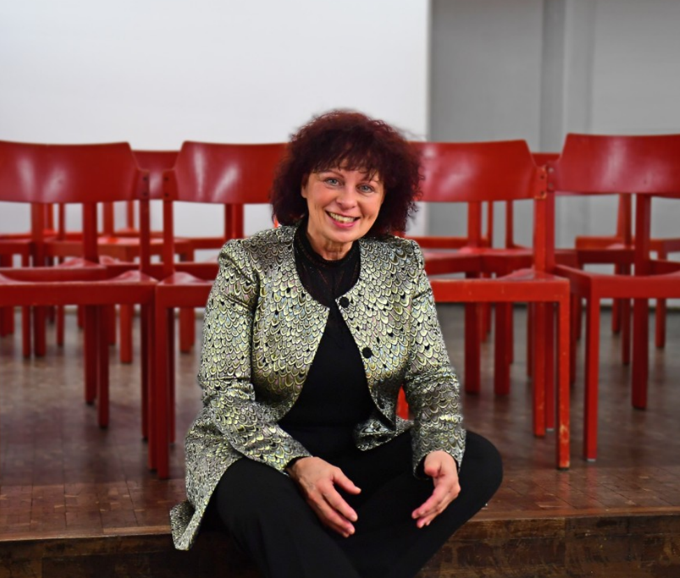 Porträt von Yvonne Sterzenbach, lächelnd auf einer Bühne vor roten Stühlen sitzend